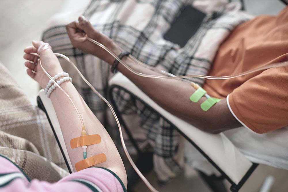 de armen van een patiënt met witte huidskleur en een patiënt met zwarte huidskleur zijn zichtbaar, ze hebben beiden een infuusnaald in de arm