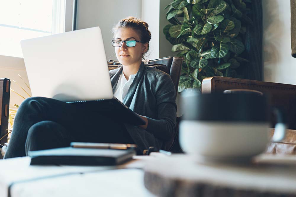 Een vrouw met bril zit in een gemakkelijke stoel met een open laptop op haar schoot. In de voorgrond staat een kopje op tafel.